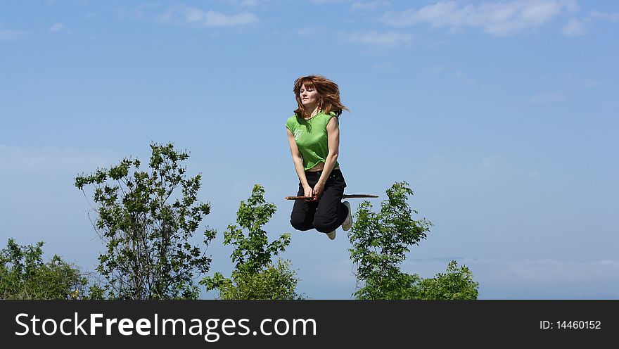 A young witch flies on a stick above trees.