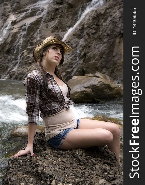 A young woman cowgirl sitting on a big rock by the river with waterfalls in the background. A young woman cowgirl sitting on a big rock by the river with waterfalls in the background.