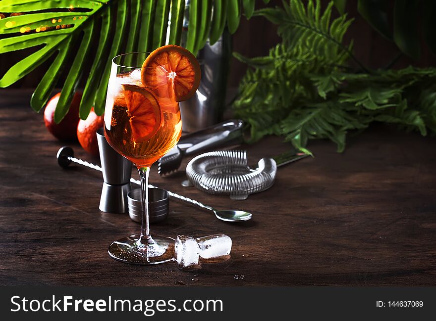 Aperol spritz cocktail in big wine glass with orange slices, summer cool fresh alcoholic cold beverage. Wooden bar counter background with tools, summer mood concept with palm leaves, copy space, selective focus