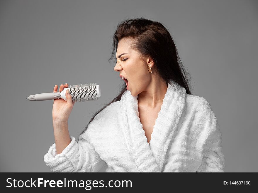 A beautiful brunette in a white terry robe with disheveled hair sings emotionally using a comb instead of a microphone. Studio photo on a gray background. A woman has a good mood in the morning. The girl is going to the party.