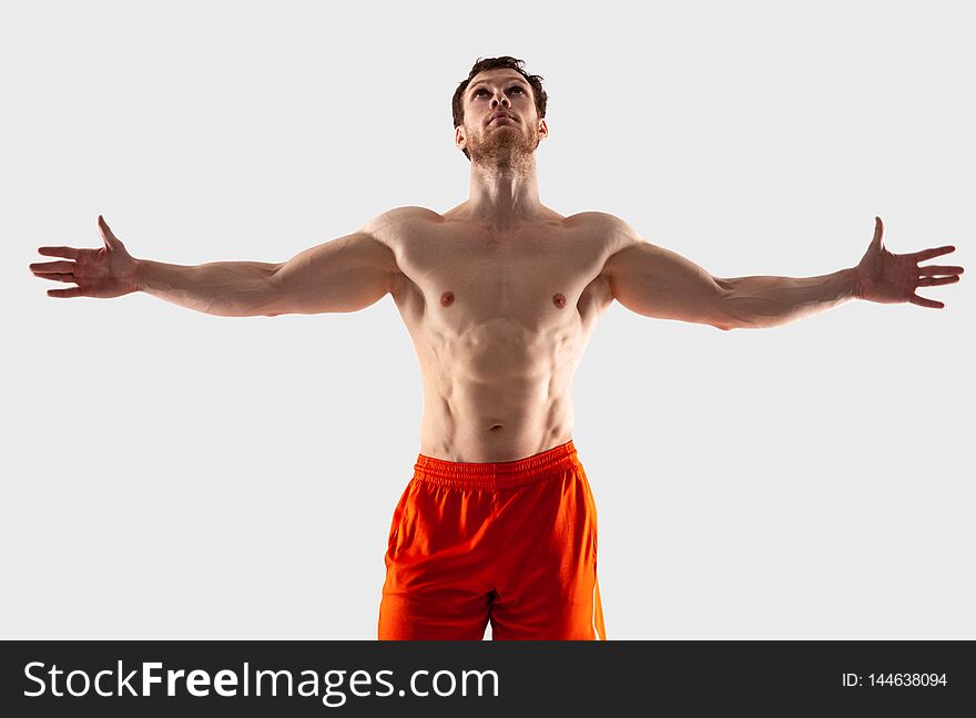 A man in orange shorts with a muscular body spread his arms to the side and looks up