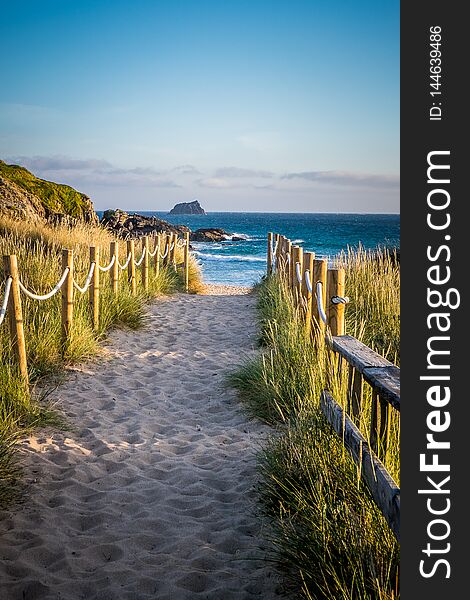 Photograph of a sandy well trodden path to the beach in Finesterre Spain. There is a rope and stick fence on each side with grasses beyond. Blue water and blue sky in the background. Photograph of a sandy well trodden path to the beach in Finesterre Spain. There is a rope and stick fence on each side with grasses beyond. Blue water and blue sky in the background.