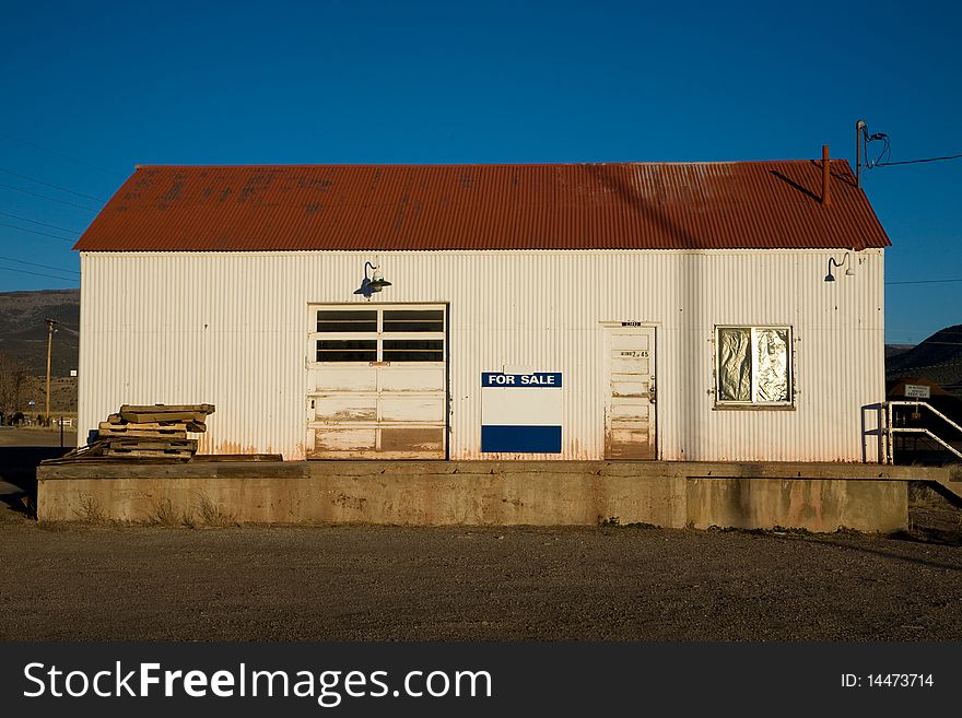 A white corrugated metal building with a red roof and a blue for sale sign stands out against a deep blue sky in the western US. A white corrugated metal building with a red roof and a blue for sale sign stands out against a deep blue sky in the western US.