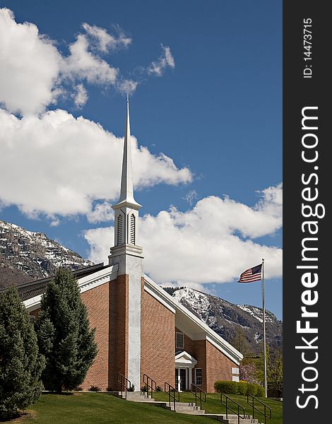 A small red brick church with a tall steeple and an American flag, with blue sky, snow-capped mountain and white clouds in the background. A small red brick church with a tall steeple and an American flag, with blue sky, snow-capped mountain and white clouds in the background