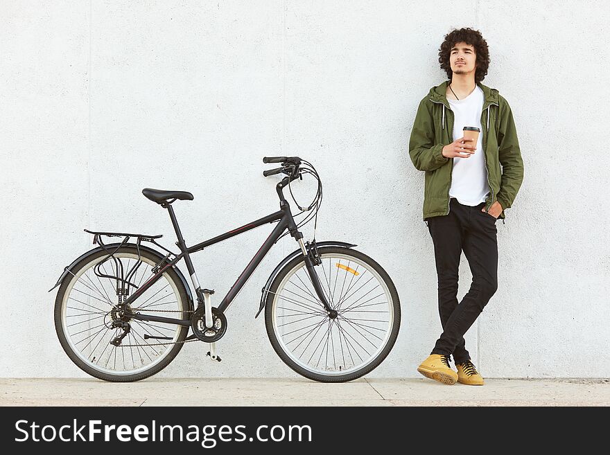 Indoor shot of attractive pencive man with crisp hair drinking hot baverage from paper cup, looks thoughtfully away, wears in fashionable clothes, stands near bicycle, isolated over white studio wall