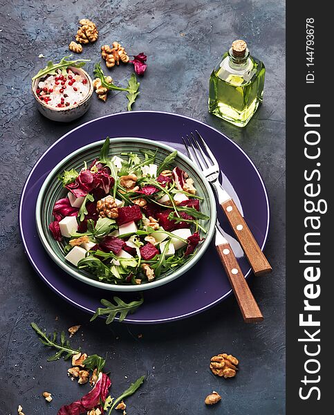 Beet or beetroot salad with fresh arugula, radicchio, soft cheese and walnuts on plate with fork, dressing and spices on blue kitchen table background, copy space, top view