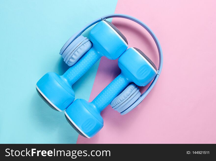 Minimalism. Blue headphones, plastic dumbbells on blue pink background. Sports with music. Flat lay. Top view