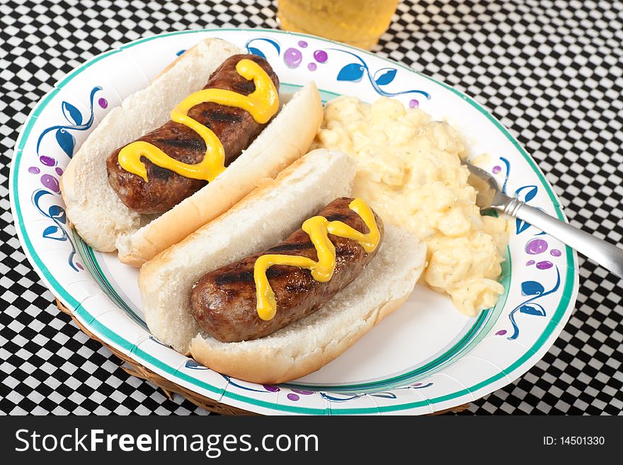 Two bratwursts on a bun with potato salad and a glass of beer on a checkered picnic table cloth. Two bratwursts on a bun with potato salad and a glass of beer on a checkered picnic table cloth