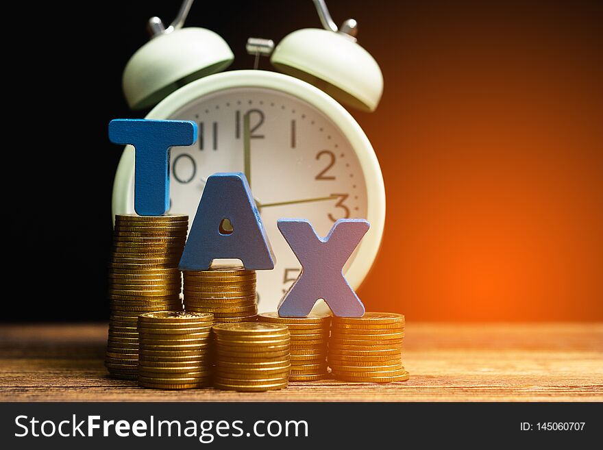 Time to pay TAX concept. TAX alphabet with stack of coin and vintage alarm clock on wooden working table in dark background