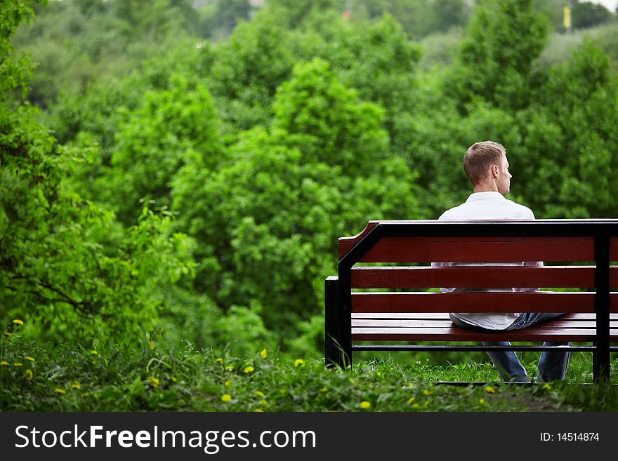 The young man sits one on a bench against green trees. The young man sits one on a bench against green trees