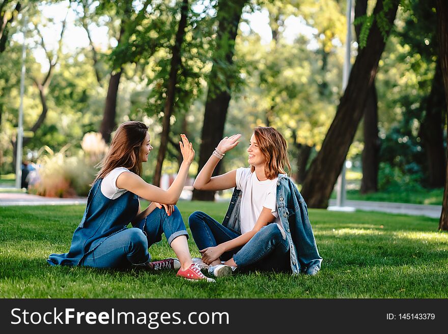 Happy female friends raising hands up giving high five sitting on green grass lawn on sunny day, outdoors in city park. Happy female friends raising hands up giving high five sitting on green grass lawn on sunny day, outdoors in city park