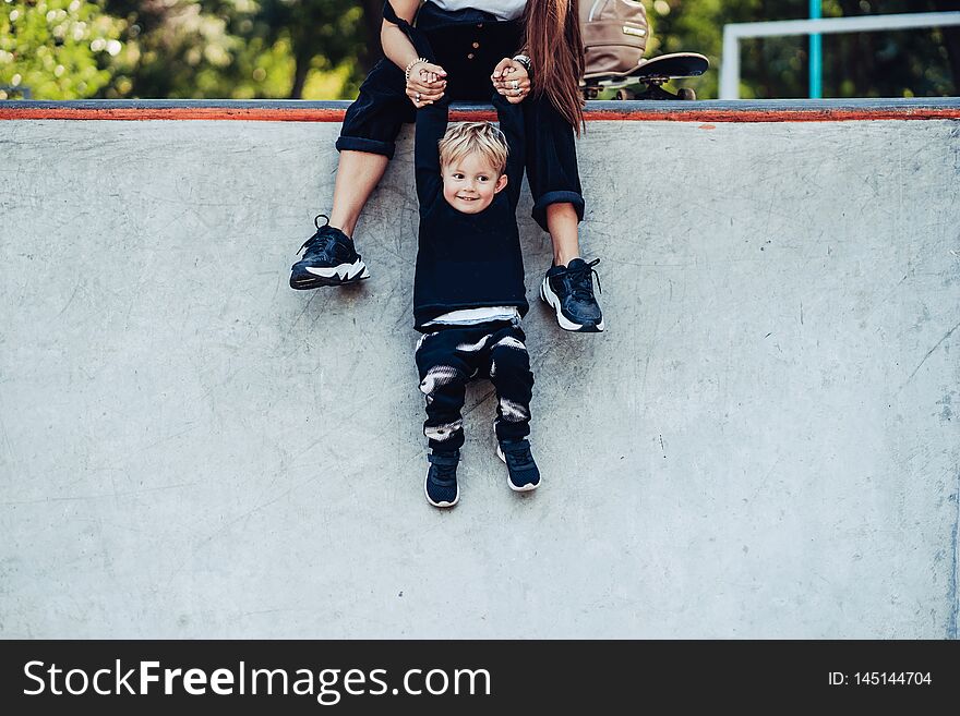 Beautiful young hipster mom holds her little son by the hands have fun at the skatepark