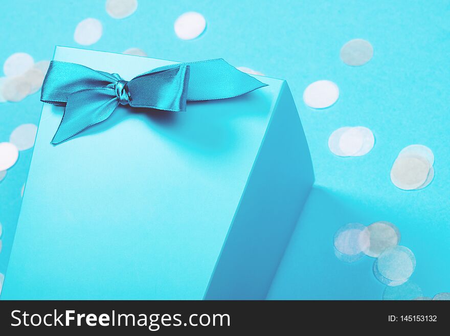 Fragment of blue box with shiny satin ribbon tied in a bow with scattered pastel confetti on the background of the same tone. Trendy creative celebration backdrop with copy space for text. Fragment of blue box with shiny satin ribbon tied in a bow with scattered pastel confetti on the background of the same tone. Trendy creative celebration backdrop with copy space for text