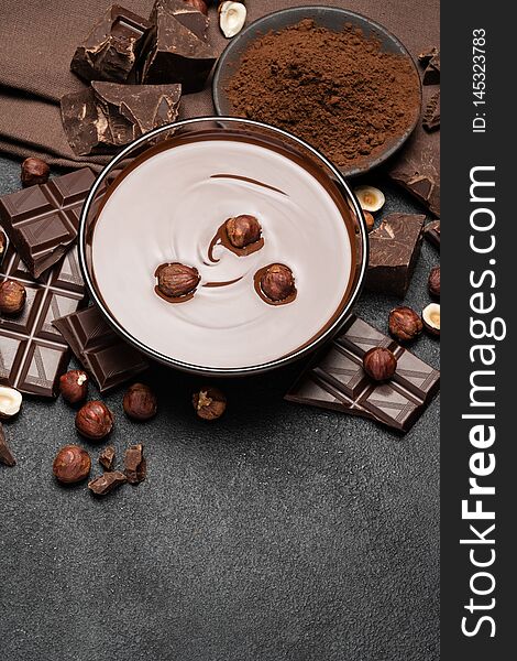 Glass bowl of chocolate cream or melted chocolate, pieces of chocolate and hazelnuts on dark concrete background