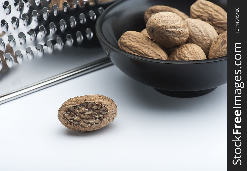 A Part Grated Nutmeg, In Front Of A Bowl Of Nutmegs and A Grater, On A White Background