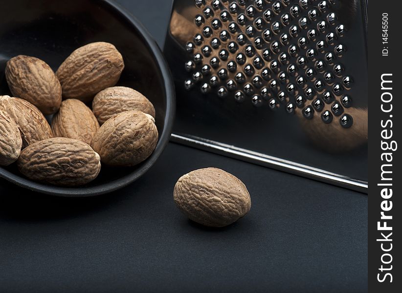 A Nutmeg On A Black Background, In Front Of A Black Dish Of Nutmegs and A Chrome Grater