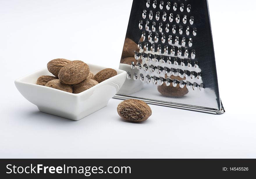 A Partly Grated Nutmeg In Front Of A Chrome Grater and Next To A White Dish Full Of Nutmegs, On A White Background