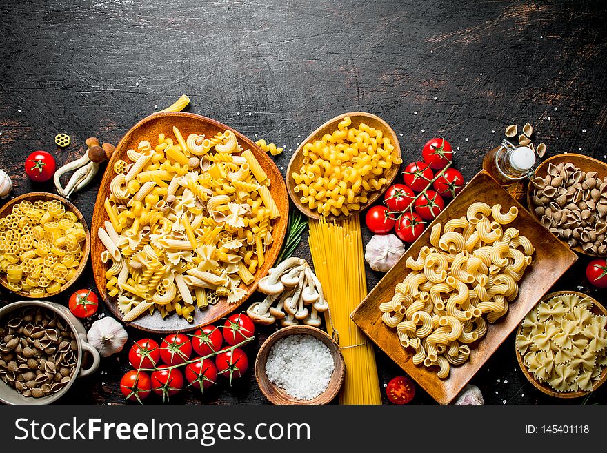 Assortment of different types of raw pasta with mushrooms, tomatoes and garlic. On dark rustic background