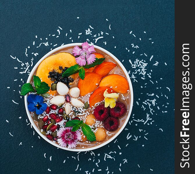 Isolated shot of bowl with tropical fruit decorated with flowers, mint, coconut flakes isolated over dark blue background. Fresh persimone, raspberry and almond