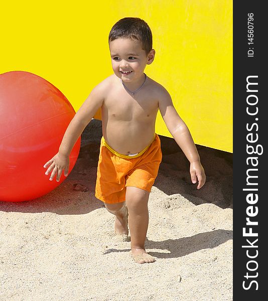 A happy little boy in an an orange swim suit, running surrounded by a huge red ball , yellow background and sand. A happy little boy in an an orange swim suit, running surrounded by a huge red ball , yellow background and sand.