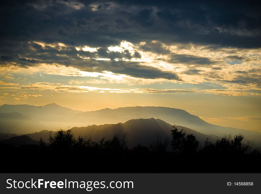 The sun sets behind storm clouds over a mountain range, casting rays of visible light and a golden glow. The sun sets behind storm clouds over a mountain range, casting rays of visible light and a golden glow.