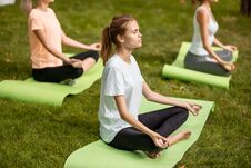 Three Young Slim Girls Sit In The Lotus Positions With Closing Eyes Doing Yoga On Yoga Mats On Green Grass In The Park Stock Photo