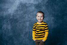 A Little Boy Poses In Front Of A Gray-blue Concrete Wall. Portrait Of A Smiling Child Dressed In A Black And Yellow Striped Royalty Free Stock Photos