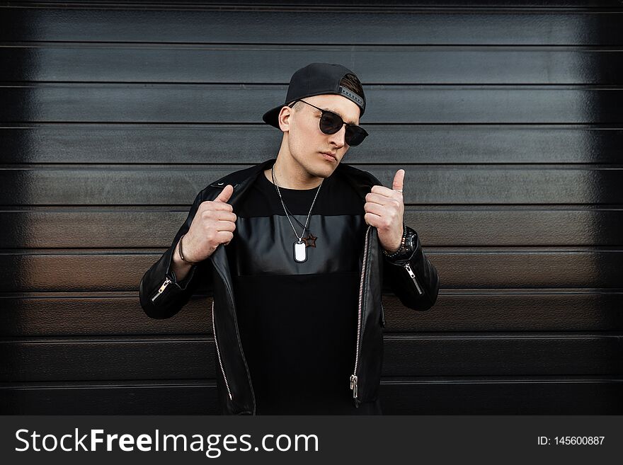 Serious young hipster man in a black baseball cap in stylish dark sunglasses in a vintage leather jacket with a fashionable t-shirt stands near a black wooden house. Handsome urban guy outdoors