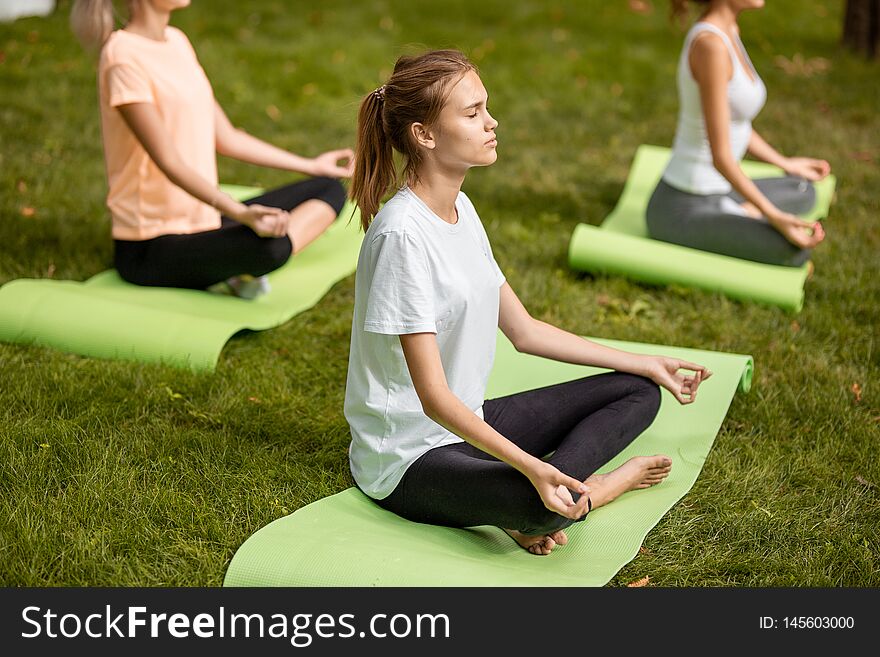 Three young slim girls sit in the lotus positions with closing eyes doing yoga on yoga mats on green grass in the park on a warm day .