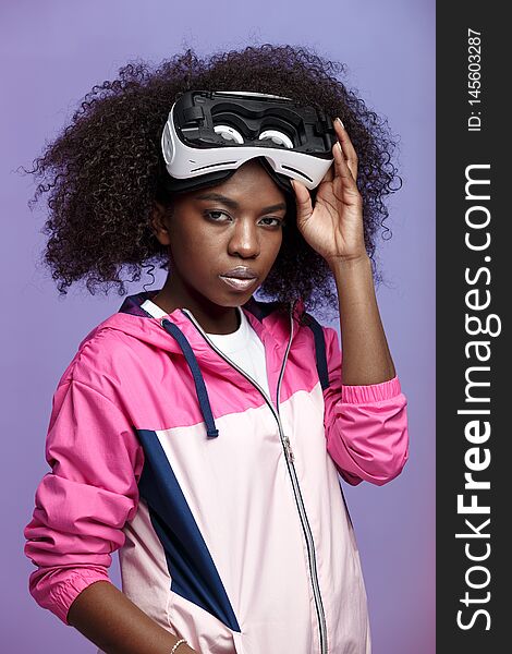 Mod Curly Brown-haired Girl Dressed In The Pink Sports Jacket Wearing On Her Head The Virtual Reality Glasses Poses In