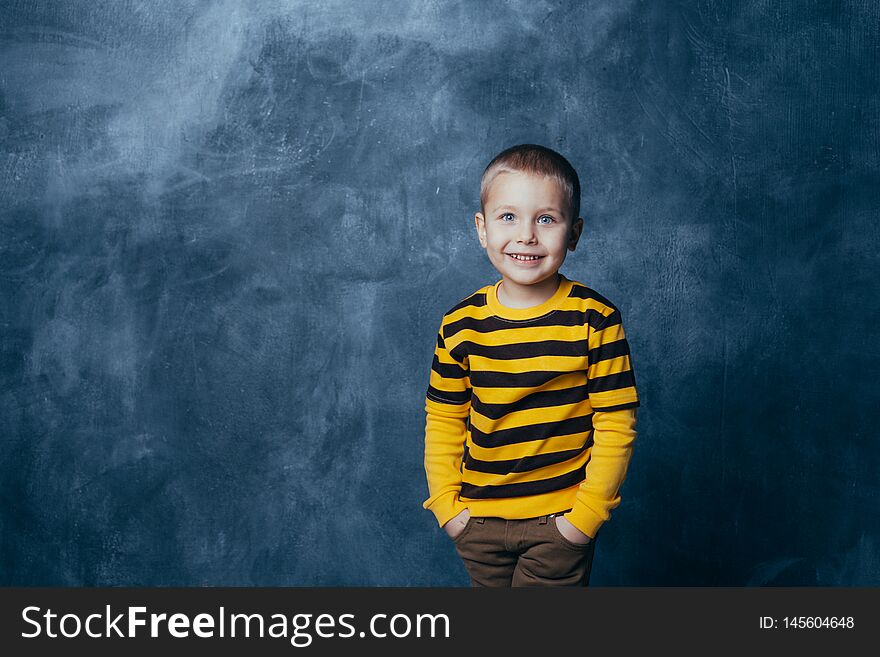 Little fashionable boy posing in front of a gray-blue concrete wall. Portrait of a smiling child dressed in a black and yellow striped sweater and brown pants. Little fashionable boy posing in front of a gray-blue concrete wall. Portrait of a smiling child dressed in a black and yellow striped sweater and brown pants