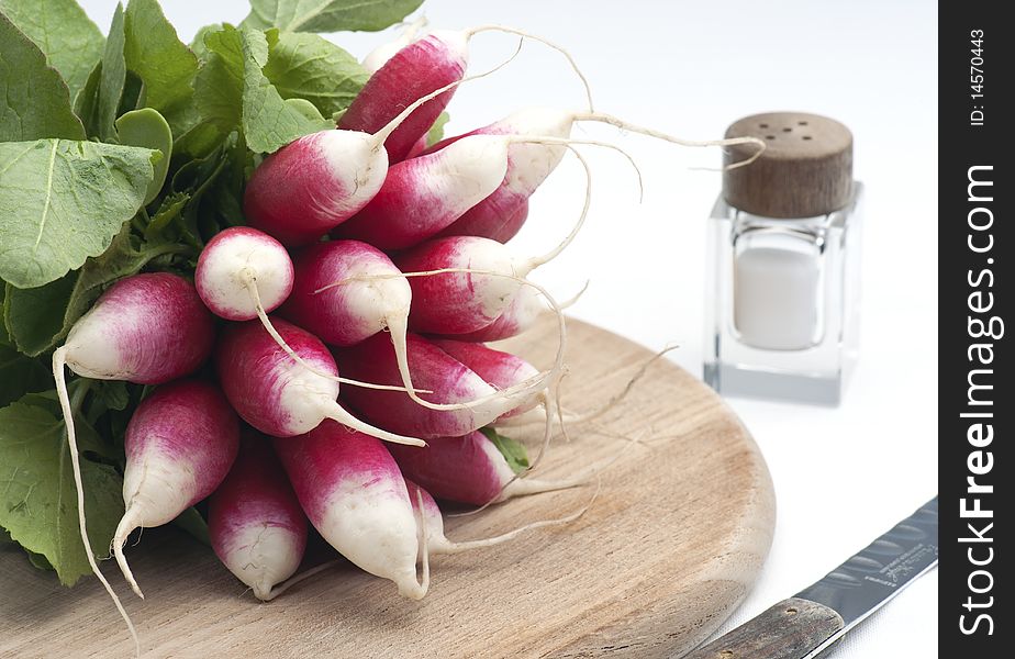 A Bunch Of Fresh French Breakfast Radish On A Wooden Chopping Board, With A Salt Pot and knife, On A White Background. A Bunch Of Fresh French Breakfast Radish On A Wooden Chopping Board, With A Salt Pot and knife, On A White Background