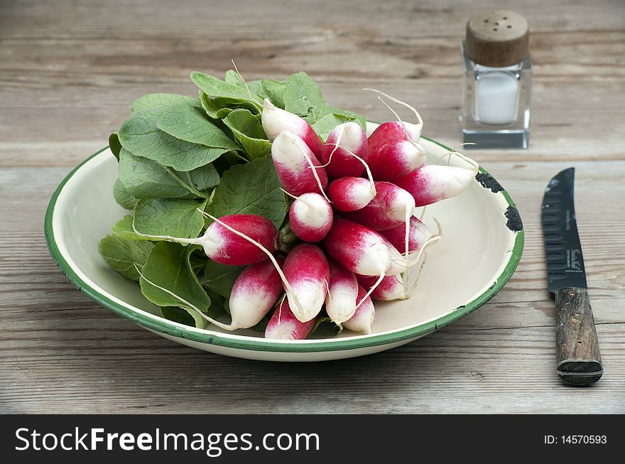 A Bunch Of Fresh French Breakfast Radish In A Enamel Dish, With A Knife and Salt Pot On A Wooden Kitchen Table. A Bunch Of Fresh French Breakfast Radish In A Enamel Dish, With A Knife and Salt Pot On A Wooden Kitchen Table
