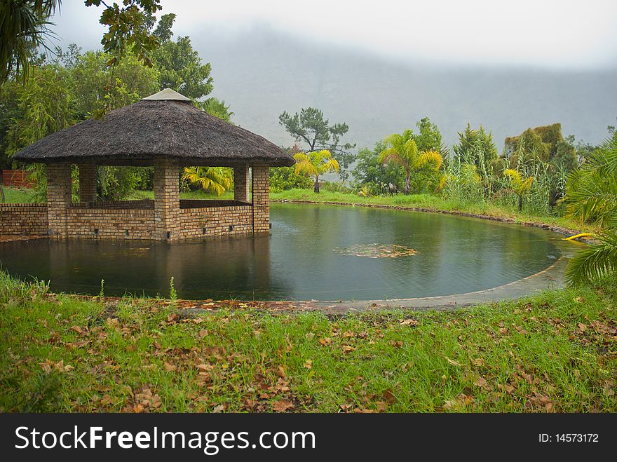 Grass roofed, brick gazebo, surrounded by a pond and grass, with storm clouds in the background