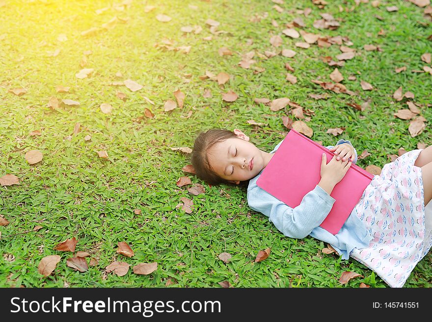 Little Girl Resting With Book Lying On Green Grass With Dried Leaves In The Summer Garden