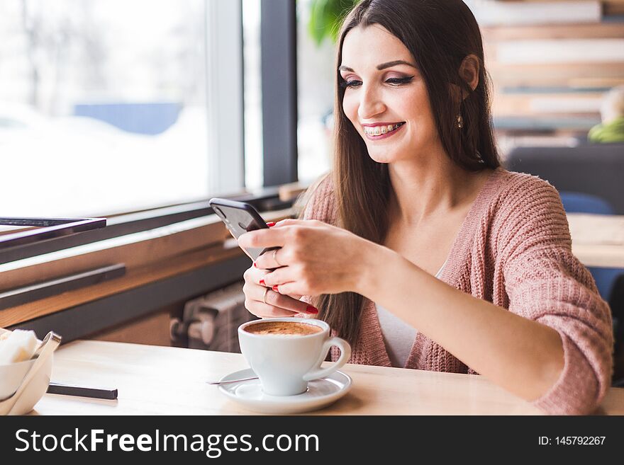 Beautiful young woman sits at a table in a cafe with a cup of coffee and uses the phone