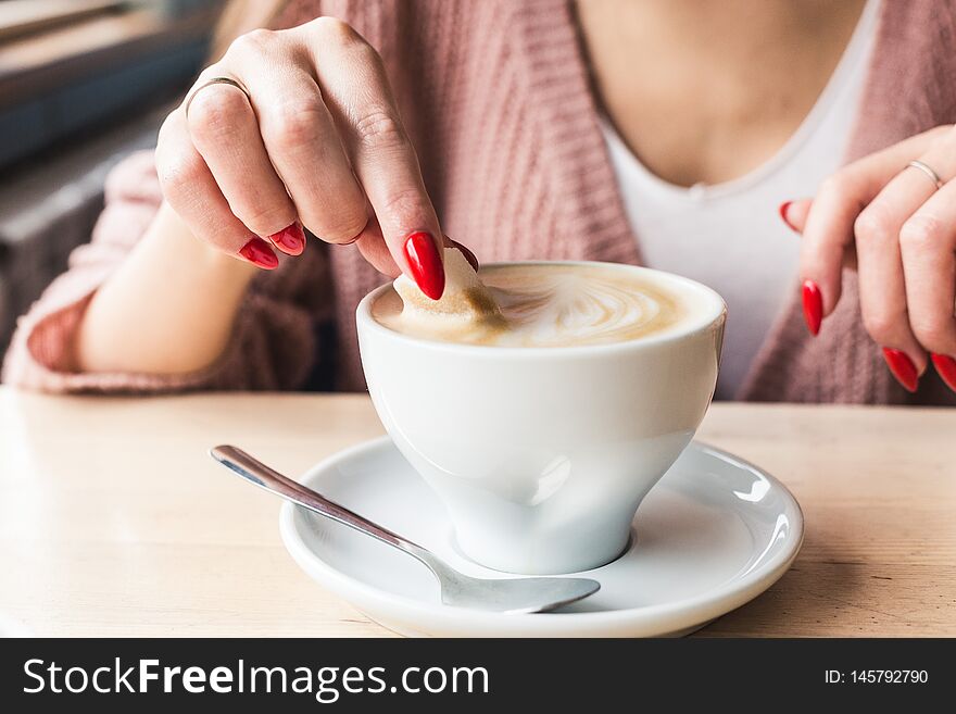 A girl dunks a piece of sugar in coffee. Closeup of hands with red nails make-up and cappuccino cup