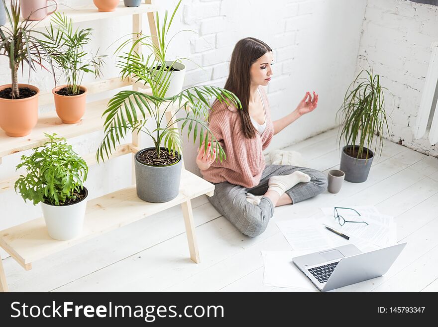 Young happy woman sits on the floor in a yoga pose in a bright apartment and works behind a laptop, a freelancer girl meditates while working. Stress of work.