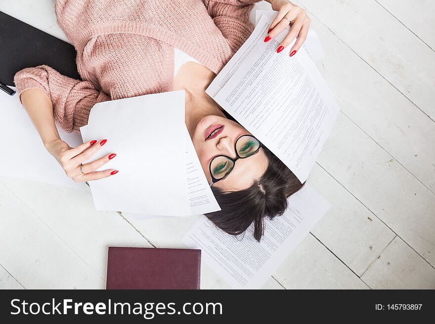 A beautiful woman in stylish glasses lies on the floor among the papers and documents, the girl freelancer smiles and relaxes during a break from work.