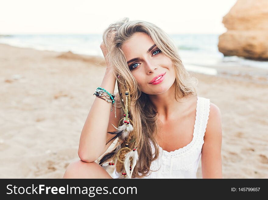 Closeup portrait of attractive blonde girl with long hair and blue eyes sitting on beach. She wears white dress