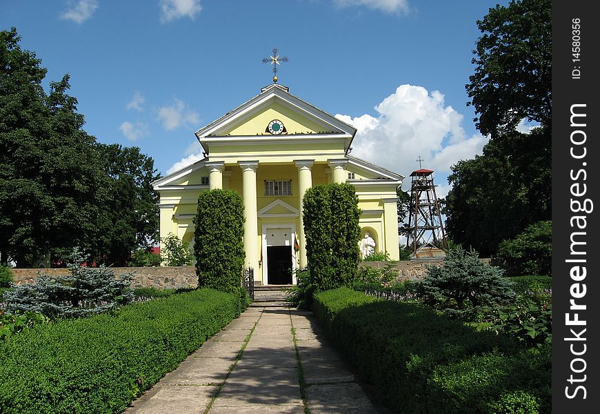 A beutiful nice yellow church with trees bushes around it.