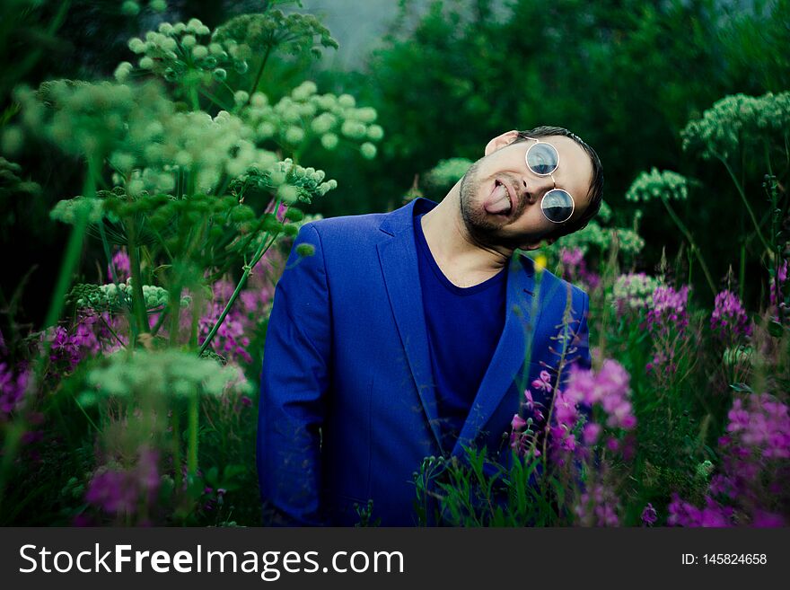 A man in a blue jacket on a flower meadow. He`s wearing blue glasses. In the meadow of purple flowers and large hats of white high flowers.