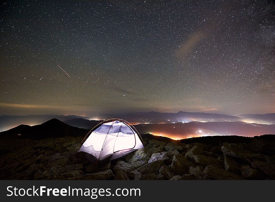 Tourist summer camping at night on the top of rocky mountain. Glowing tent under incredibly beautiful night sky full of stars. On background amazing starry sky, mountains and luminous villages. Tourist summer camping at night on the top of rocky mountain. Glowing tent under incredibly beautiful night sky full of stars. On background amazing starry sky, mountains and luminous villages