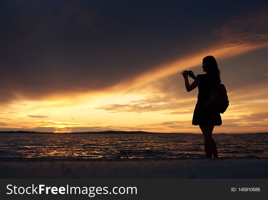 Silhouette of slim woman in short dress and with backpack and camera standing alone at water edge taking picture of beautiful seascape at sunset. Tourism and vacations concept. Silhouette of slim woman in short dress and with backpack and camera standing alone at water edge taking picture of beautiful seascape at sunset. Tourism and vacations concept.