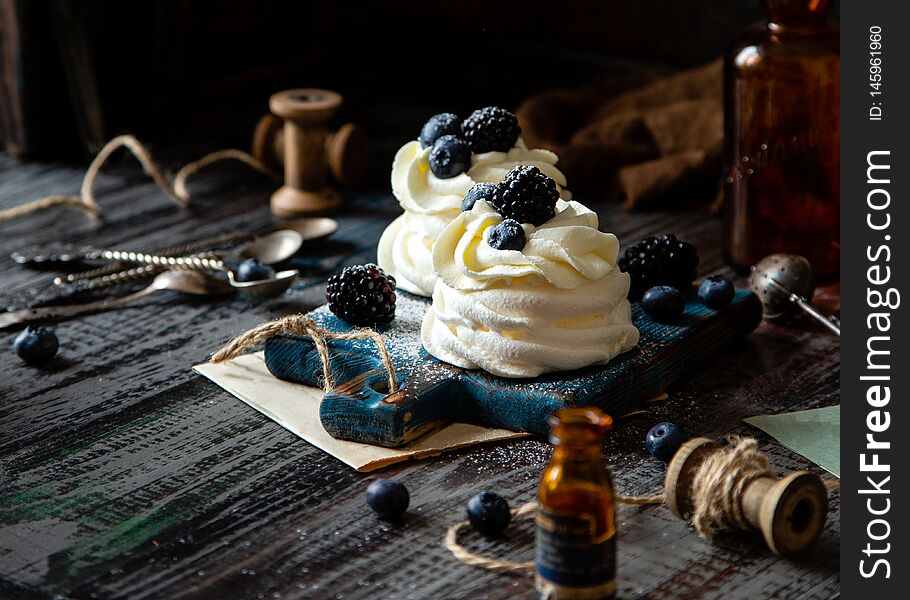 Two homemade white mini desserts pavlova on wooden blue board with whipped cream and blueberries, blackberries on rustic table with berries, old bottles, spoons, spool of thread, brown towel