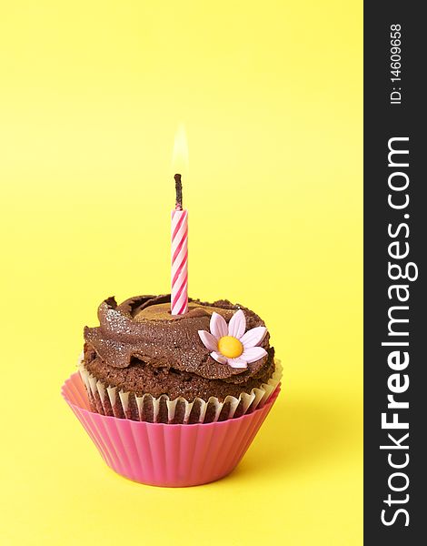 Miniature chocolate cupcake with icing, decorative flower and birthday candle on yellow background. Miniature chocolate cupcake with icing, decorative flower and birthday candle on yellow background