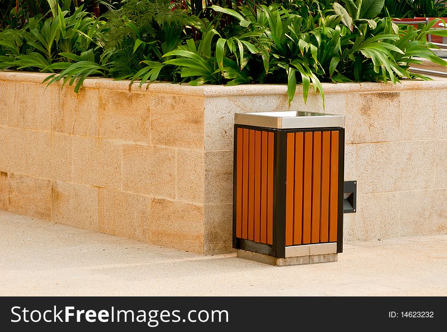 A Modern Wooden Slatted Litter Bin in a Park with Granite and Plant Background. A Modern Wooden Slatted Litter Bin in a Park with Granite and Plant Background