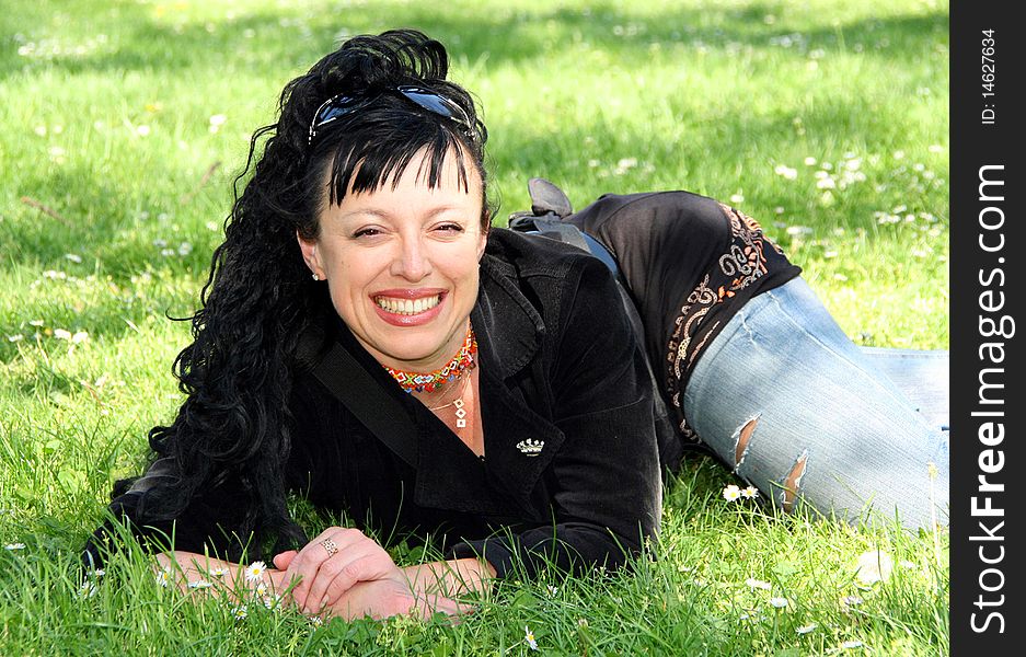 A beautiful woman in a black jacket, fragmentary jeans and with sun glasses on a head lies on a grass