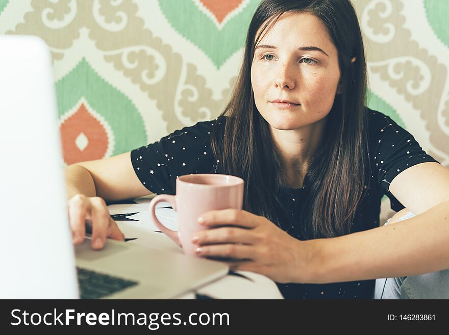 Female freelancer looks at a laptop monitor and holds a cup of coffee, business concept. Female freelancer looks at a laptop monitor and holds a cup of coffee, business concept