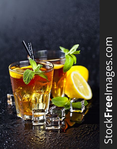 Two glasses with cold traditional iced tea with lemon, mint leaves and ice cubes on wet black background